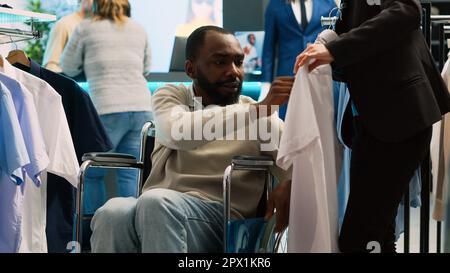 Kunde mit Behinderung besucht einen modernen Bekleidungsladen und möchte trendige Freizeitkleidung im Einkaufszentrum kaufen. Junger Mann im Rollstuhl, der modische Kleidung an Kleiderbügeln untersucht. Handgeführte Aufnahme. Stockfoto