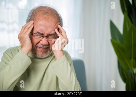 Kopfschmerzen. Trauriger asiatischer Seniorenmann sitzt auf dem Sofa und fühlt sich verletzt und einsam, ältere Menschen halten Kopf mit Hand leiden an Migräne Kopfschmerzen, Gesundheit im Alter Stockfoto