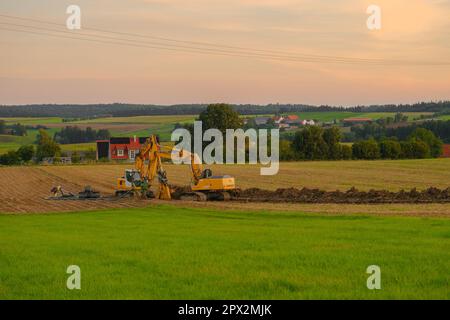 Rohrverlegung. Bagger auf dem Feld im Dorfhintergrund. Bau von Maschinen und Feldarbeiten. Durchführung von Kommunikationen Stockfoto