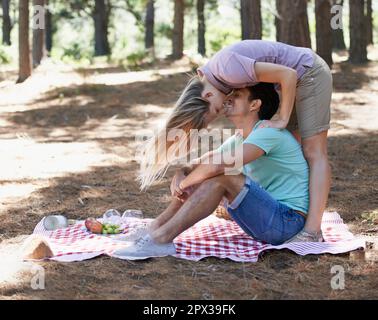 Verspielte Romantik. Ein junger Mann, der draußen auf einer Picknickdecke sitzt, mit seiner Freundin, die sich für einen Kuss über ihn lehnt. Stockfoto