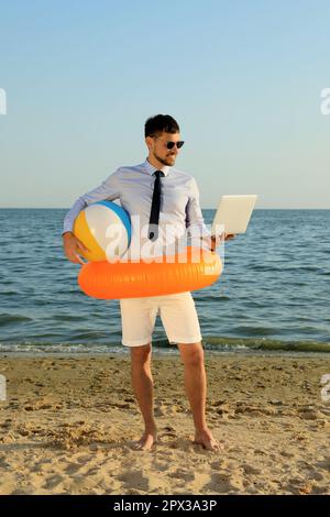 Glücklicher Mann mit aufblasbarem Ring, Ball und Laptop in der Nähe des Meeres am Strand. Geschäftsreise Stockfoto