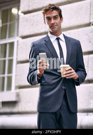 Guten Morgen. Ein gutaussehender Geschäftsmann in einem Anzug, der einen Kaffee zum Mitnehmen und sein Handy in der Hand hat. Stockfoto