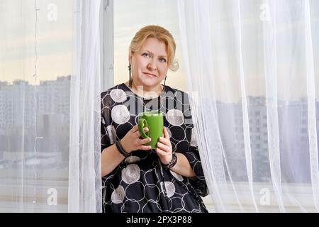 Eine fröhliche Frau mittleren Alters sitzt auf der Fensterbank und hält eine Tasse mit heißem Kakao Stockfoto
