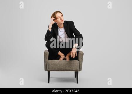 Junger Barfußmann im schwarzen Anzug, der im Sessel im Studio sitzt Stockfoto