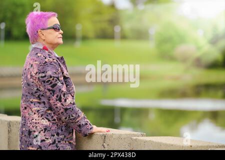 Lustige ältere Frau mit kurzen rosafarbenen Haaren und Wollmantel, die am Flussufer im Stadtpark steht Stockfoto