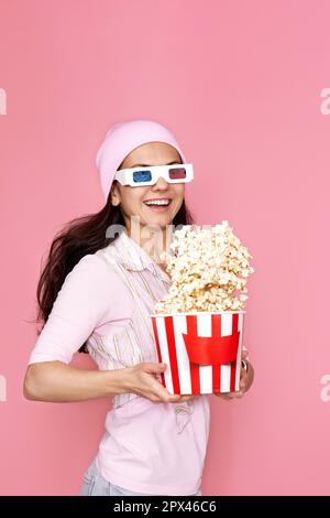 Fröhliche, glückliche Frau in 3D-Brillen, Hut mit Eimer Popcorn und Springen isoliert auf pastellrosa Hintergrund. Stockfoto