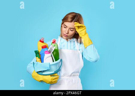 Müde, blonde Frau in gelben Handschuhen, die einen Eimer mit Reinigungsmittel auf blauem Hintergrund hält. Stockfoto