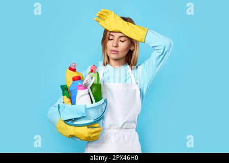 Müde blonde kaukasische Frau in Handschuhen hält Eimer mit Reinigungsmittel auf blauem Hintergrund. Stockfoto