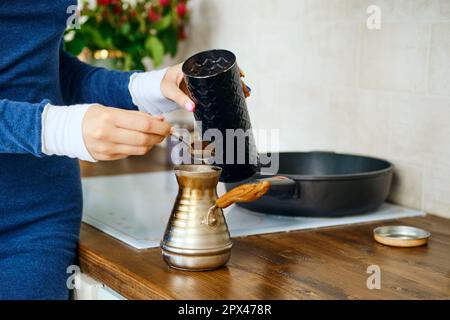 Nahaufnahme der weiblichen Hand mit Löffel mit gemahlenem Kaffee in der Nähe eines türkens. Kaffee in einer Metalltopf zubereiten. Stockfoto