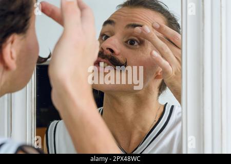 Reflexion im Spiegel einer jungen, nicht binären Latino-Person, die in ihrem Zimmer steht und mit dem Finger auf den Augenlidern Make-up aufträgt. Stockfoto