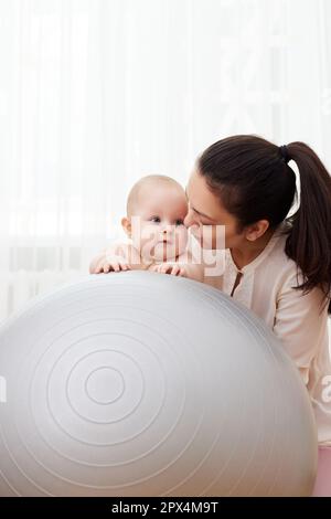 Glückliche Mutter, die zu Hause mit ihrem kleinen Mädchen auf einem Fitnessball spielt Stockfoto