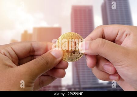 Blick auf die Hand, die eine Bitmünze hält und sie anderen Personen über die Gebäude im Stadthintergrund gibt. Das Konzept des Kryptowährungshandels Stockfoto