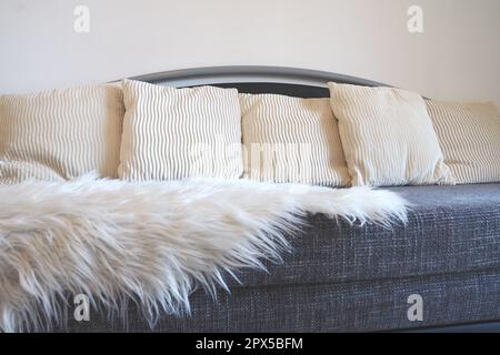 Graues Sofa mit grobem Polstertuch und weißen dekorativen, gewellten Kissen. Über den Sitz wird ein weißer Kunstpelzteppich oder ein Überwurf geschleudert Stockfoto