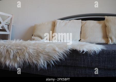 Graues Sofa mit Polsterbezug und weißen, dekorativen Kissen. Weißer Teppich oder Bettüberwurf aus Kunstfell mit langem Stapel. Wohnzimmer ist drin Stockfoto