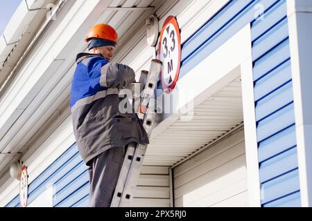 Ein Elektriker steht auf der Leiter und ersetzt die Glühlampe in der Deckenleuchte am Tor eines Industriegebäudes oder einer Garage. Echter Workflow Stockfoto