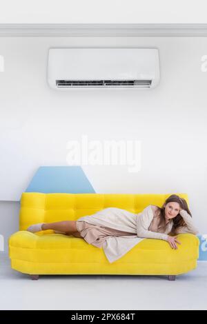 Junge, glückliche weiße Frau liegt auf einem gelben Sofa in heller Inneneinrichtung mit geteilter Klimaanlage Stockfoto