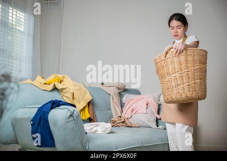 Eine wunderschöne Haushälterin, die Hausarbeit macht, hält einen Holzkorb mit sauberer, dreckiger Kleidung, um zu Hause zu waschen, eine asiatische Hausfrau hält einen Korb mit h in der Hand Stockfoto