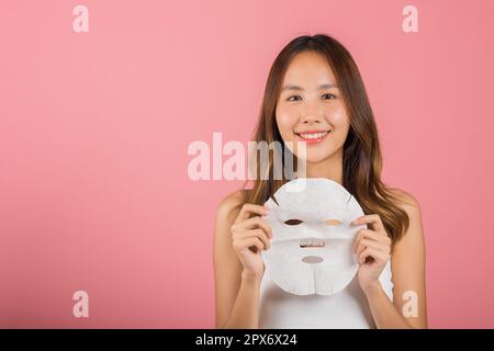 Gesundheitszentrum. Portrait schöne Frau lächelt hält Gesichtsmaske, Studio Shot isoliert auf blauem Hintergrund, asiatische Frau hält reinigende Maske Stockfoto