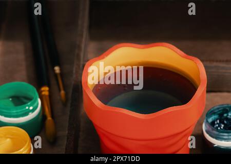 Ein Wasserbecher zum Reinigen von Pinseln, drei Gouache-Farben und zwei Pinsel in Staffelbox, Nahaufnahme. Stockfoto