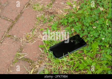 Smartphone liegt auf Gras in der Nähe von Straßenbelägen im Freien. Verloren und gefunden Stockfoto