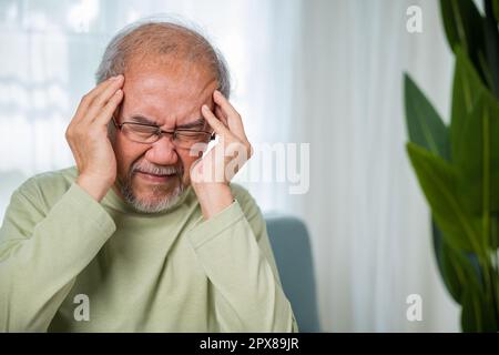 Kopfschmerzen. Trauriger asiatischer Seniorenmann sitzt auf dem Sofa und fühlt sich verletzt und einsam, ältere Menschen halten Kopf mit Hand leiden an Migräne Kopfschmerzen, Gesundheit im Alter Stockfoto