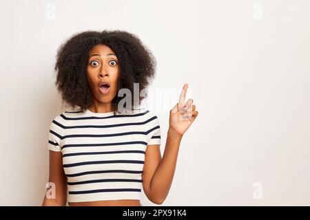 Beeindruckte junge Frau mit lockigen Haaren Zeigefinger rechts, zeigt etwas auf leerem Raum, trägt gestreiftes T-Shirt, isoliert auf weißem Hintergrund Stockfoto