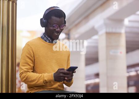 Ein lächelnder junger Mann mit kabellosen Kopfhörern kommuniziert online mit seinem Smartphone, während er in der U-Bahn steht Stockfoto