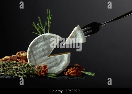 Brie-Käse mit Walnüssen, Thymian und Rosmarin auf schwarzem Hintergrund. Brie Käse und Gabel mit Dreieckstück. Stockfoto