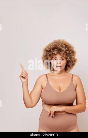 Die vertikale Aufnahme einer jungen kurvigen Frau reagiert fröhlich und zeigt nach oben auf einen Kopierbereich isoliert neben dem weißen Werbekonzept. Hochwertiges Foto Stockfoto