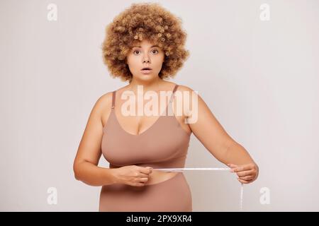 Eine ziemlich dicke Frau mit überraschten und unerwarteten Emotionen, überprüfe ihre Parameter mit einem Maßband, isoliert neben einer weißen Wand. Stockfoto