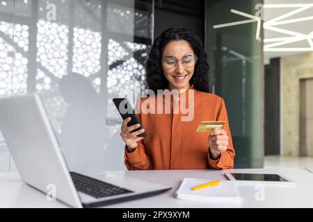 Fröhliche und erfolgreiche hispanische Geschäftsfrau, die im Büro arbeitet, Frau mit Bankkreditkarte und Telefon, Online-Shopping und erfolgreiche Buchung, Frau am Arbeitsplatz mit Laptop. Stockfoto