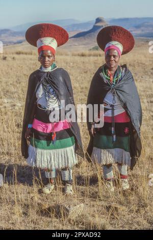 KWAZULU-NATAL, SÜDAFRIKA - 2. MAI 2023: Zwei Zulu-Frauen in traditionellen Kleidern und mit einem Iicholo-Hut vor dem Isandlwana-Hügel. Stockfoto