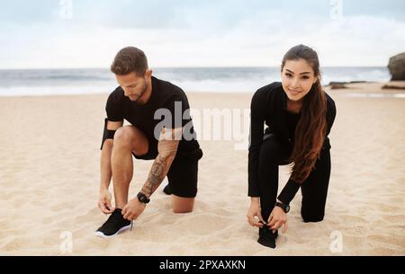 Eine junge Dame und ein Mann in Sportbekleidung binden Schuhe am Strand und bereiten sich auf das morgendliche Workout vor Stockfoto