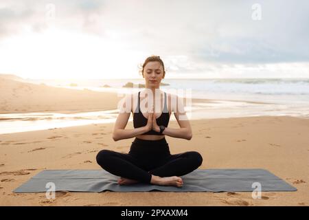 Junge ruhige Frau in Sportbekleidung, die Yoga praktiziert, mit geschlossenen Augen am Strand nahe dem Meer meditiert, Kopierraum Stockfoto