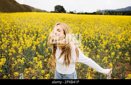 Wunderschöne, blonde, fröhliche Frau, die einen Hut trägt, posiert inmitten eines Feldes blühender gelber Rapsblumen Stockfoto