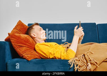 Der Mann ruht sich auf der Couch, die mit einer Decke bedeckt ist, und liest Nachrichten auf dem Smartphone Stockfoto