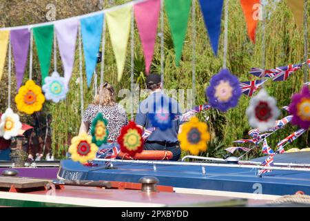 London, Vereinigtes Königreich, 29. April 2023: Die 40. Jährliche Canalway Cavalcade findet während des Mayday-Feiertagswochenendes in Little Venice in London statt. Ein Mann und eine Frau sitzen auf dem Dach eines Schmalbootes, mit Wimpel- und Häkelblütengardlands im Vordergrund und einer weinenden Weide im Hintergrund. Anna Watson/Alamy Live News. Stockfoto