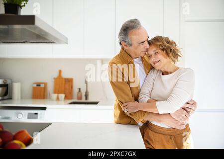 Liebevolle ältere Ehepartner, die sich umarmen, zärtliche Momente zu Hause genießen, in der Küche stehen, in der Küche, Kopierraum Stockfoto