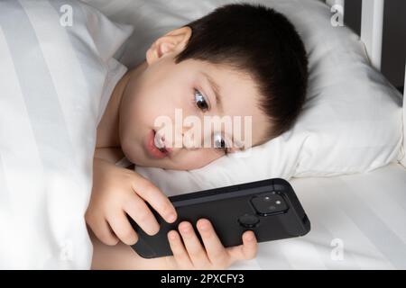 Ein 5-jähriger Junge spielt Spiele auf seinem Handy, während er im Bett liegt. Geräte im Bett vor dem Schlafengehen bei einem Kind Stockfoto