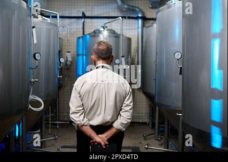 Rückblick auf den Brauereimann in der Schürze zwischen Brennereibehältern in der Bierfabrik Stockfoto