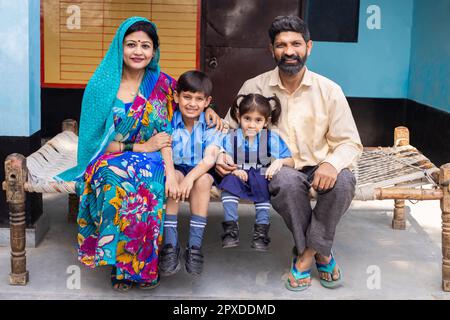 Porträt von jungen, glücklichen indischen Eltern, die mit kleinen Söhnen und Tochter in Schuluniform sitzen, ländliches indien, Bildungskonzept. Stockfoto