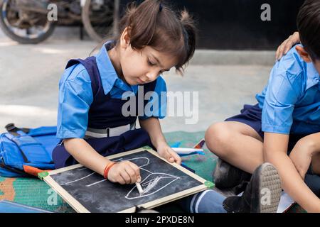 Süße kleine indische Schülerin, die Schuluniform trägt und auf Schieferbrett mit Kreide schreibt, während sie auf dem Boden sitzt. Regierungsschule. Ländliches indien. Stockfoto