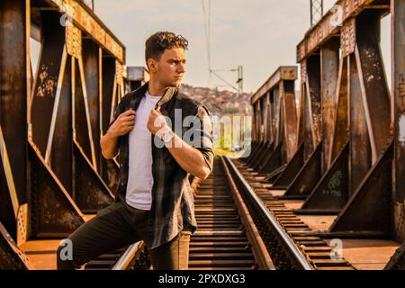 Kaukasischer Teenager, der auf Bahngleisen auf einer rostigen Eisenbahnbrücke steht, mit einer verschwommenen Stadt im Hintergrund Stockfoto