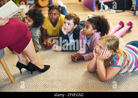 Wir lieben gute Geschichten. Eine vielfältige Gruppe von Kindern, die auf dem Boden des Klassenzimmers liegen und ihrem Lehrer beim Lesen eines Buches zuhören. Stockfoto
