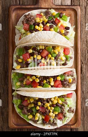 Mehltortillas gefüllt mit Hackfleisch und Gemüse, Salat, Tomaten, Mais, grüner Paprika und Bohnen, serviert auf Holzbrett, fotografierte Over hea Stockfoto