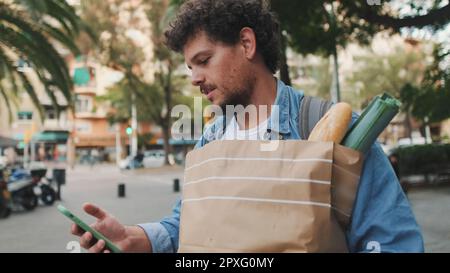 Lächelnder junger Mann in Jeansshirt mit Papiertüte und gesundem Essen in den Händen geht die Straße entlang, surft auf dem Smartphone in sozialen Netzwerken Stockfoto