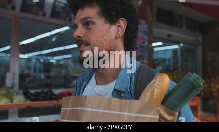 Glücklicher Mann in Jeansshirt mit Papiertüte mit gesundem Essen in den Händen geht vorbei am Öko-Markt, schaut auf die Theke Stockfoto