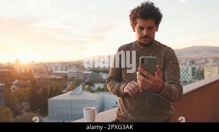 Ein lächelnder Typ steht morgens auf dem Balkon und macht Selfie auf dem Smartphone Stockfoto