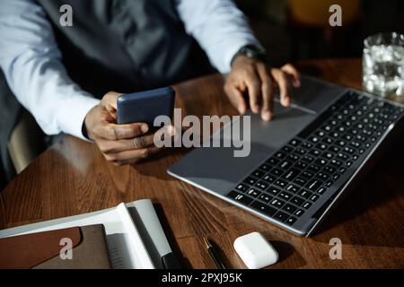 Nahaufnahme eines jungen Geschäftsmanns, der sein Smartphone mit dem Laptop verbindet und online arbeitet, während er im Café am Tisch sitzt Stockfoto