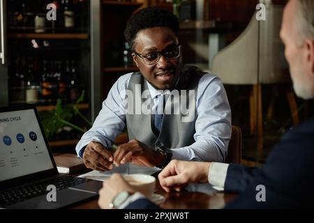 Ein afroamerikanischer Geschäftsmann, der mit seinem Kollegen spricht, während er an einem Laptop arbeitet, während er sich im Restaurant trifft Stockfoto